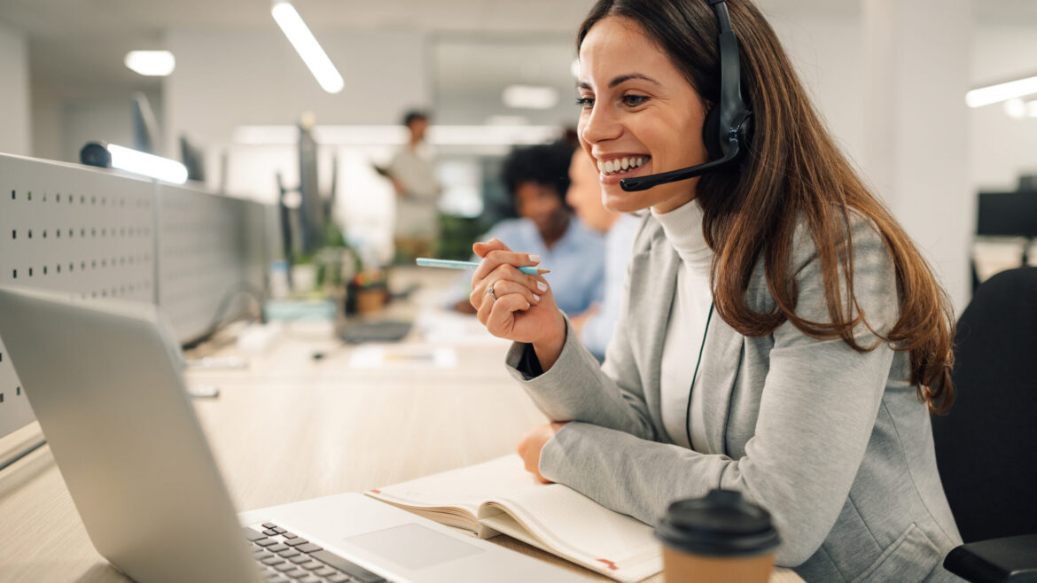 Photo of a woman, smiling with a headset on, looking at the laptop