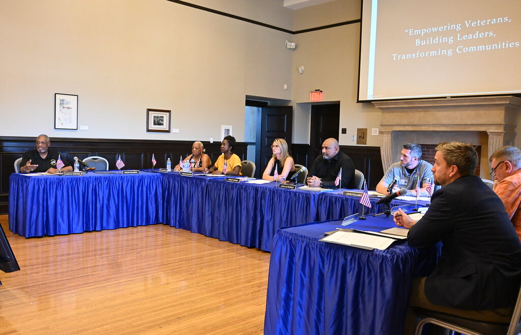 Veterans Advisory Commission meeting in session at Bob Hope Patriotic Hall. Commissioners sit around tables covered in blue cloth with small U.S. flags, discussing veteran programs. A screen in the background displays the words 'Empowering Veterans, Building Leaders, Transforming Communities.
