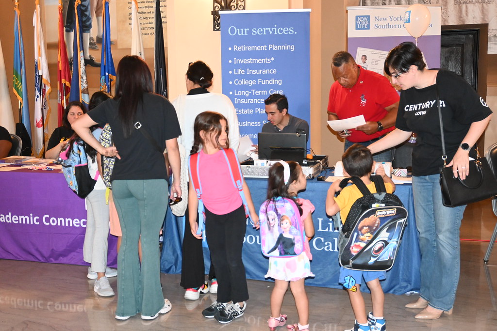 A group of children wearing backpacks stand in line with adults at an information table during a community event. Volunteers behind the table share resources on financial planning and services.