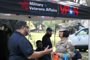 Leo Chirinos speaks with U.S. Army Reservist Spc. Jou Chi who is in uniform under an MVA and VPAN tent at a stand down event with other attendees and resource booths visible in the background.