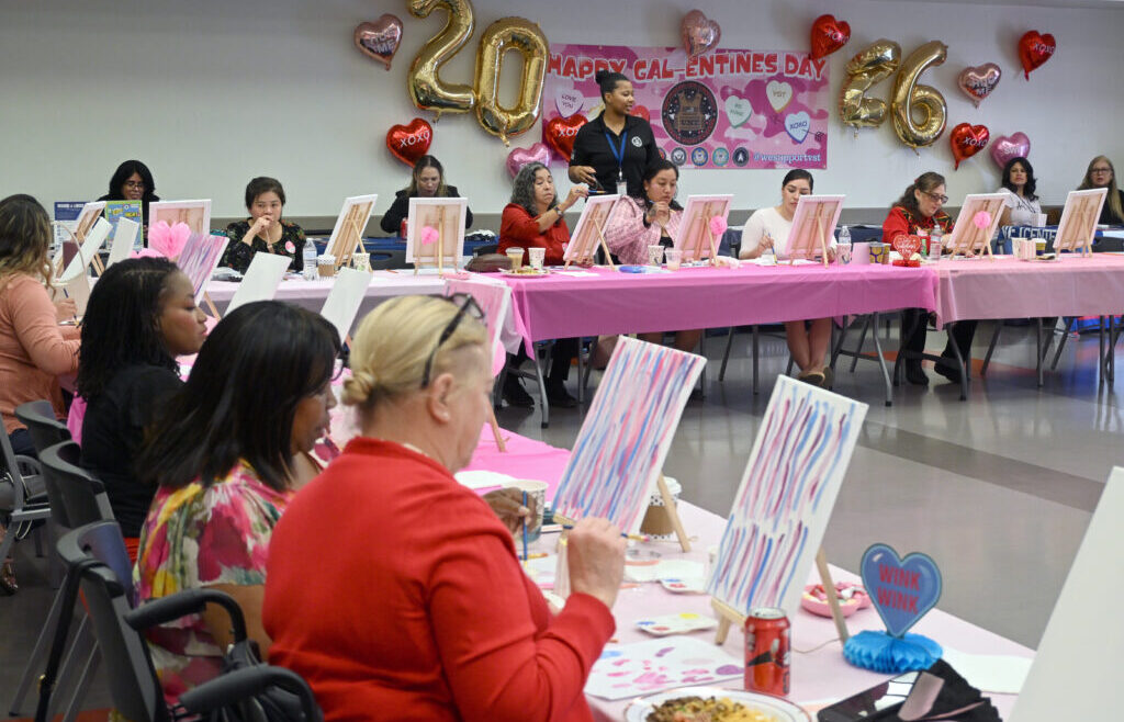 Women veterans sit at long tables covered in pink tablecloths, painting on canvases during a Galentine&rsquo;s Sip n&rsquo; Paint event. Valentine&rsquo;s Day decorations, including heart balloons and a &ldquo;Happy Galentine&rsquo;s Day&rdquo; banner with large gold &ldquo;2026&rdquo; balloons, hang on the wall behind them as an organizer stands assisting participants.