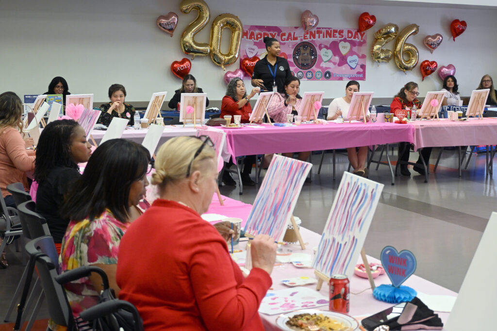 Women veterans sit at long tables covered in pink tablecloths, painting on canvases during a Galentine&rsquo;s Sip n&rsquo; Paint event. Valentine&rsquo;s Day decorations, including heart balloons and a &ldquo;Happy Galentine&rsquo;s Day&rdquo; banner with large gold &ldquo;2026&rdquo; balloons, hang on the wall behind them as an organizer stands assisting participants.