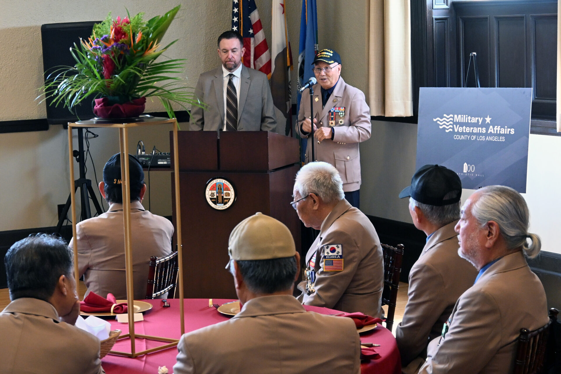 Vietnam War veteran speaks at a podium during a luncheon at Bob Hope Patriotic Hall, as fellow veterans seated at round tables listen, with U.S. flags and a Los Angeles County Military and Veterans Affairs display in the background.