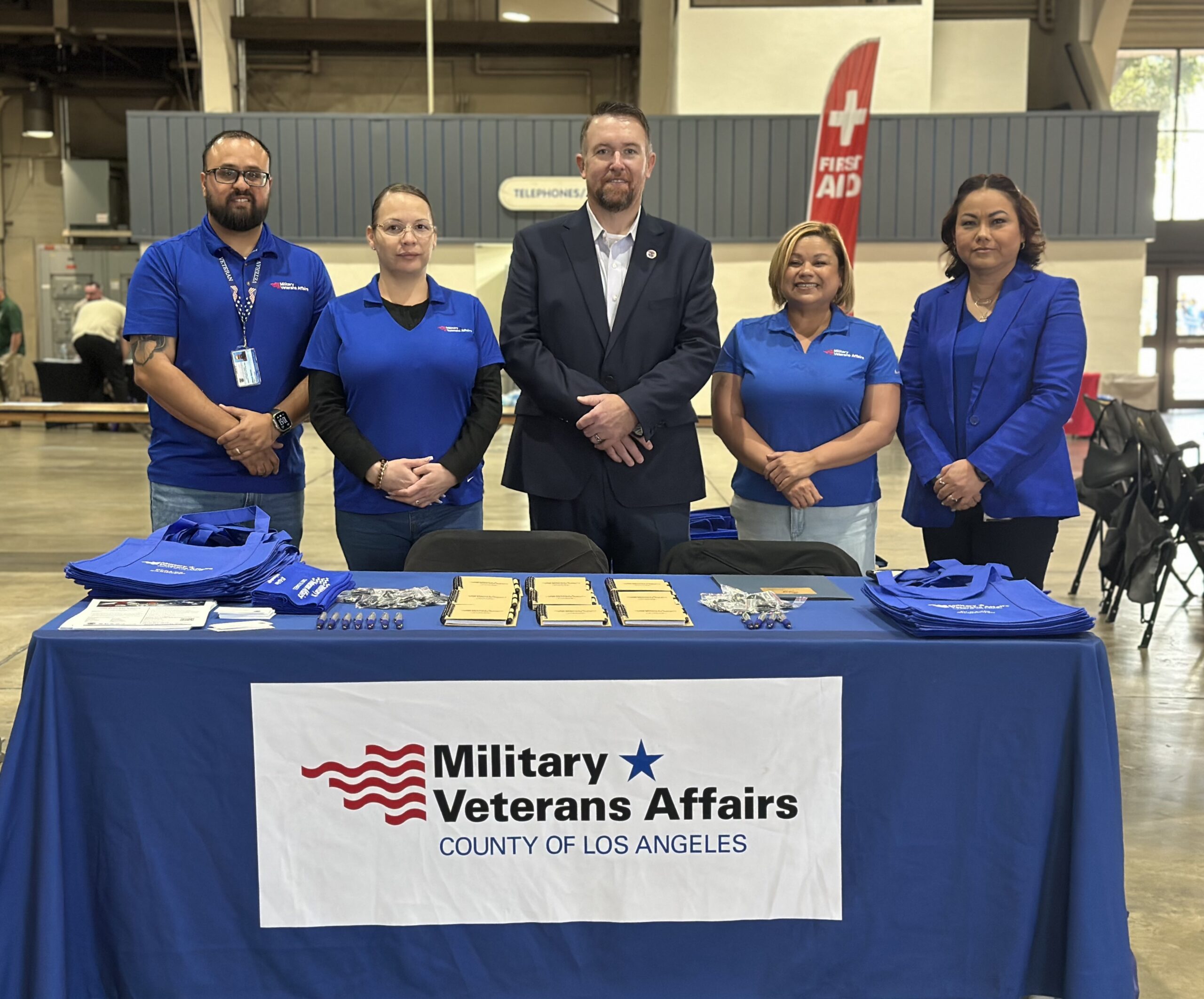 Five Los Angeles County Military and Veterans Affairs representatives stand behind an outreach table with branded materials at an indoor event.