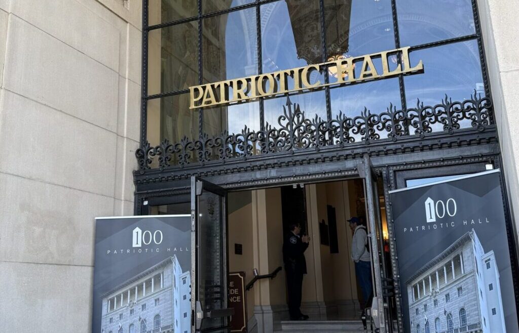 Entrance of Bob Hope Patriotic Hall with ornate ironwork and gold &ldquo;Patriotic Hall&rdquo; lettering above the doors, flanked by two large banners featuring the building and &ldquo;100 Patriotic Hall,&rdquo; marking the Centennial celebration.