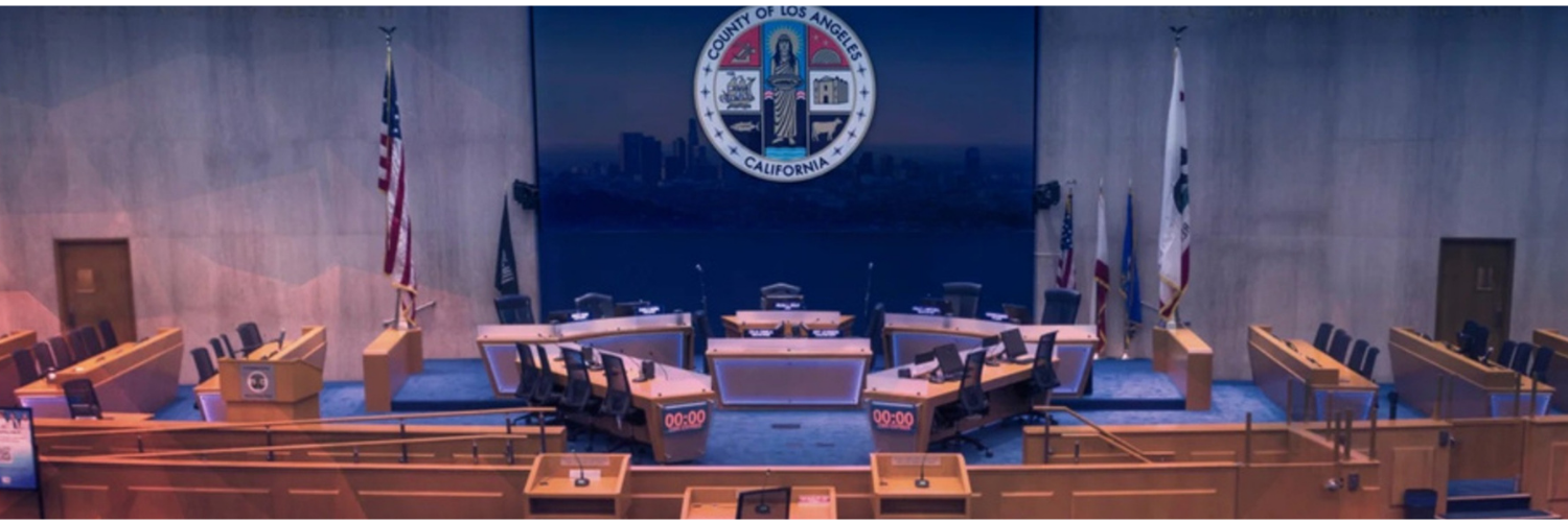 Los Angeles County Board of Supervisors chamber with seating, podium, and county seal displayed at the front.