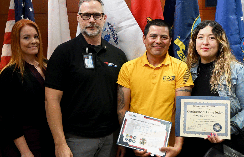 Group in a courtroom stands with a Veterans Treatment Court graduate holding certificates, with U.S. and military flags and the Los Angeles County seal in the background.
