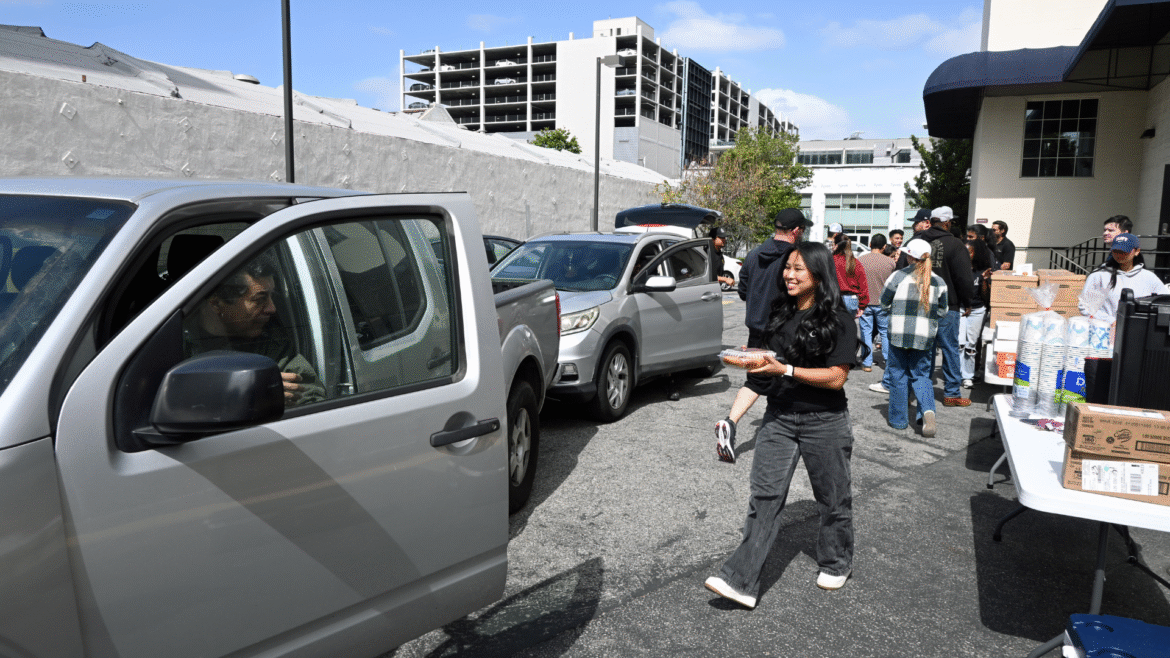 Volunteers distribute food and drinks to people in cars during a drive-through event, handing items through car windows while others gather near tables with supplies in a parking lot.