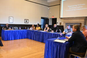 Veterans Advisory Commission meeting in session at Bob Hope Patriotic Hall. Commissioners sit around tables covered in blue cloth with small U.S. flags, discussing veteran programs. A screen in the background displays the words 'Empowering Veterans, Building Leaders, Transforming Communities.