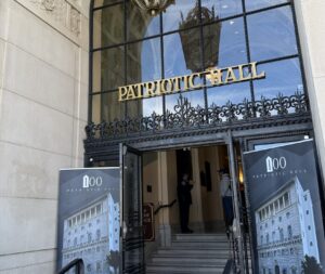 Entrance of Bob Hope Patriotic Hall with ornate ironwork and gold “Patriotic Hall” lettering above the doors, flanked by two large banners featuring the building and “100 Patriotic Hall,” marking the Centennial celebration.
