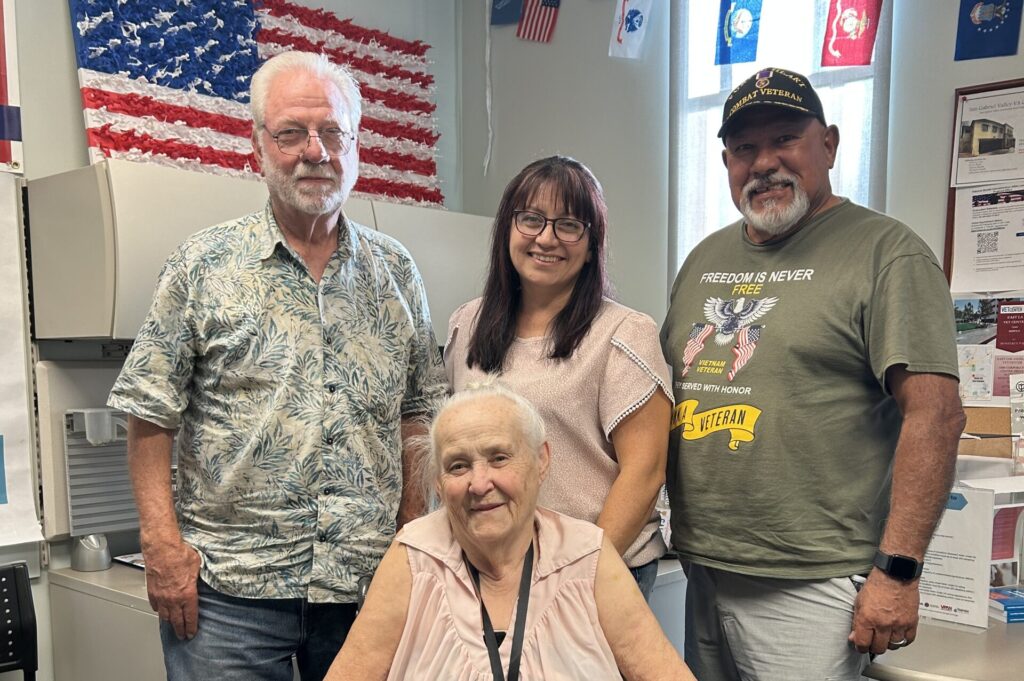 Four people gather inside a decorated office space with military flags and an American flag made of red, white, and blue materials displayed on the wall. An older woman in a wheelchair sits at the front, smiling, wearing a light pink blouse and black-and-white patterned pants. Behind her stand two men and a woman—one man in a green military veteran shirt and cap, another in a patterned button-up shirt, and a woman in glasses and a light pink top. The group appears friendly and proud, surrounded by veteran-related displays.