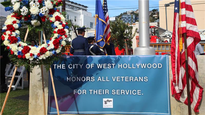 A Veterans Day ceremony in West Hollywood featuring a large red, white, and blue floral wreath, uniformed honor guard members holding American flags, and a banner reading ‘The City of West Hollywood honors all veterans for their service.